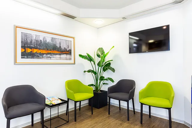 Wartezimmer mit zwei grünen und zwei grauen Stühlen, einem kleinen Tisch mit Zeitschriften, einer großen Pflanze und einem an der Wand hängenden Fernseher sowie einem gerahmten Bild mit orangefarbenen Akzenten